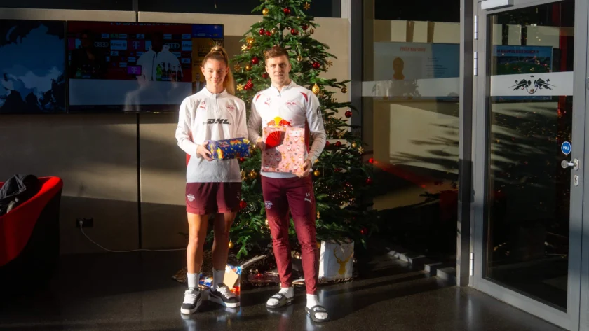 Zwei Personen in Sportkleidung stehen vor einem geschmückten Weihnachtsbaum und halten Geschenke in den Händen. Im Hintergrund sind Fernsehbildschirme und eine Tür mit einem "Push"-Schild zu sehen.