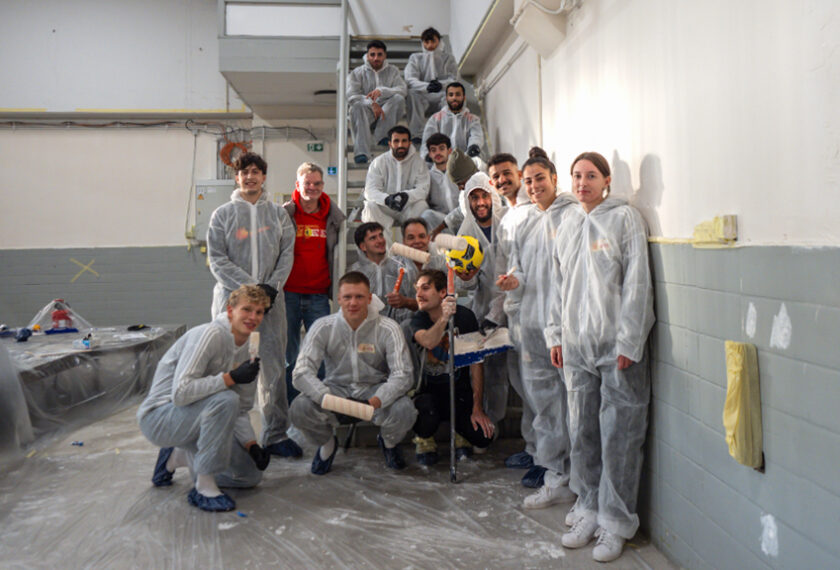 Eine Gruppe von Menschen in weißen Schutzanzügen steht und kniet in einem Raum mit grauen Wänden und einem Treppenaufgang im Hintergrund. Sie halten Farbrollen und Pinsel und lächeln in die Kamera. Einige von ihnen halten einen gelben Fußball. Der Boden ist mit Plastikfolie ausgelegt, und es gibt Anzeichen von Renovierungsarbeiten.