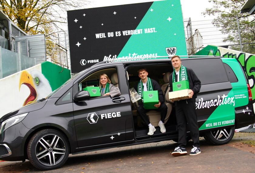 Drei Personen stehen vor einem schwarzen Lieferwagen mit grüner und weißer Weihnachtsdekoration. Sie halten grüne Geschenkboxen in den Händen und lächeln in die Kamera. Der Lieferwagen hat die Aufschrift "Weihnachtsexpress" und "Weil du es verdient hast" sowie das Logo eines Fußballvereins. Im Hintergrund ist ein Zaun mit Bäumen zu sehen.