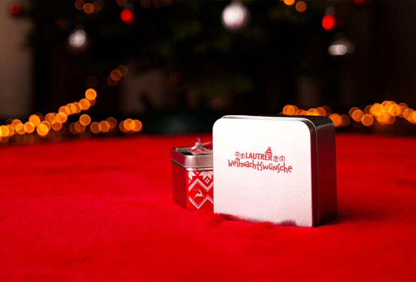 Zwei Geschenkboxen auf rotem Samt mit weihnachtlicher Beleuchtung im Hintergrund. Die größere Box zeigt den Text 'Lautrer Weihnachtswünsche' mit einem Fußball- und Weihnachtsbaum-Emoji.