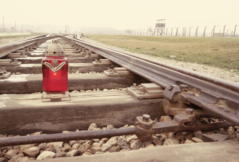 Eine rote Kerze steht auf einem Gleis im Konzentrationslager Auschwitz-Birkenau. Im Hintergrund sind Stacheldrahtzäune und Wachtürme zu sehen, die in nebligem Wetter nur schemenhaft erkennbar sind.