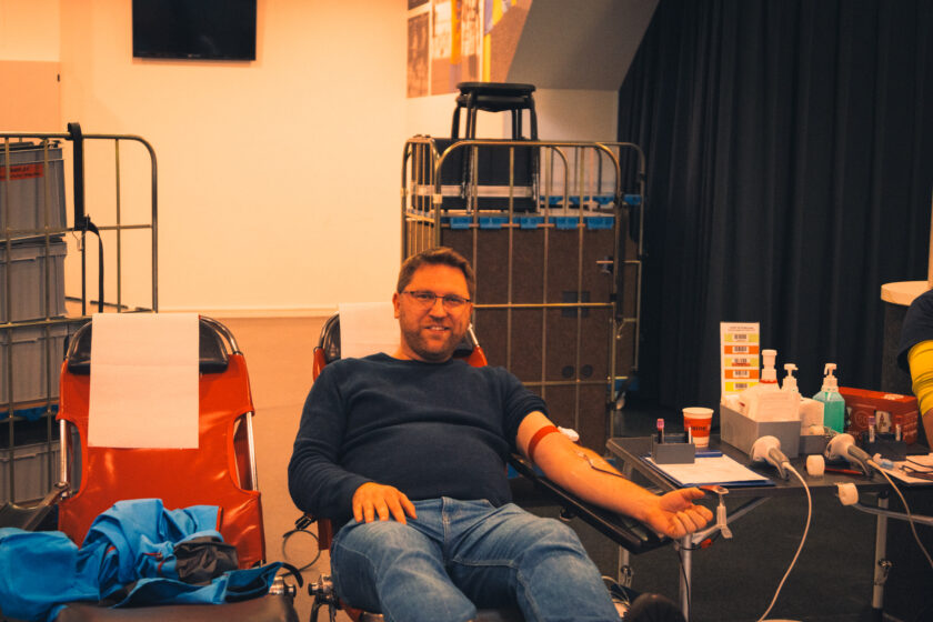 Ein Mann sitzt entspannt in einem roten Sessel und spendet Blut. Er trägt eine schwarze Brille, ein schwarzes Langarm-Shirt und blaue Jeans. Im Hintergrund sind medizinische Geräte und Vorräte zu sehen, darunter ein Tisch mit Desinfektionsmittel, Handschuhen und anderen Utensilien.