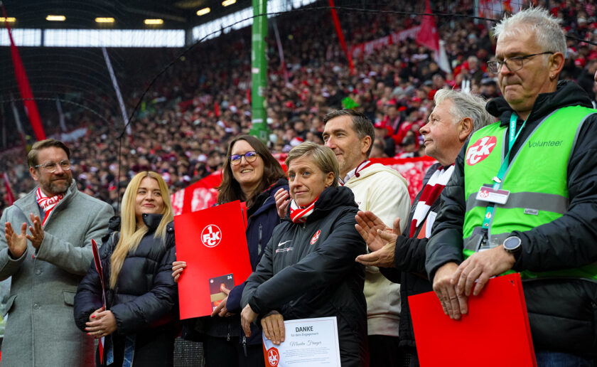 Gruppe von Menschen im Stadion, die für ihren ehrenamtlichen Einsatz gedankt wird. Sie halten rote Schilder mit dem Logo des 1. FC Köln und eine Dankeskarte. Im Hintergrund sind viele Zuschauer zu sehen.