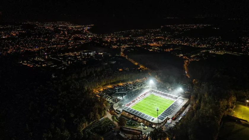 Ein Fußballstadion bei Nacht, beleuchtet und umgeben von einem dunklen Wald und einer beleuchteten Stadt im Hintergrund.
