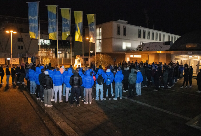 Eine Gruppe von Menschen in blauen Jacken steht nachts vor einem Gebäude und beobachtet eine Kerze, die auf einem Sockel platziert ist. Im Hintergrund sind mehrere Flaggen mit blauen und gelben Streifen zu sehen. Die Szene wirkt feierlich und erinnert an eine Gedenkveranstaltung.