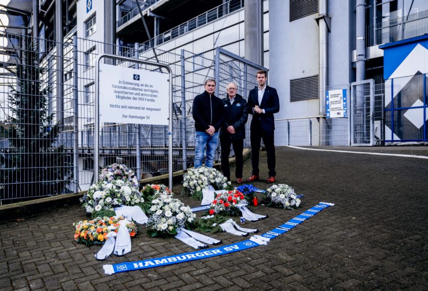 Drei Männer stehen vor einem Stadion und gedenken der Opfer des Nationalsozialismus, die der HSV-Familie angehörten. Vor ihnen liegen mehrere Kränze und Blumenarrangements auf dem Boden, darunter auch ein blau-weißer Kranz mit dem Logo des Hamburger SV. Ein Schild mit einer Inschrift erinnert an die deportierten und ermordeten Mitglieder der HSV-Familie. Die Männer haben die Hände hinter dem Rücken verschränkt und blicken ernst.