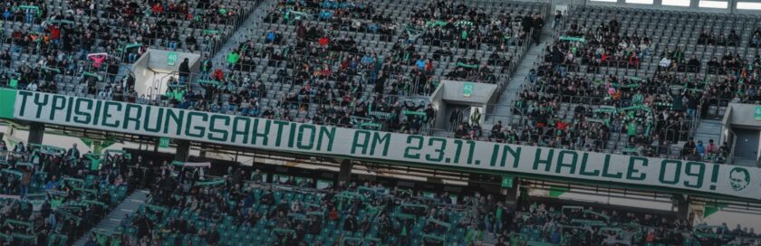 Ein volles Fußballstadion mit vielen Zuschauern, die grüne Schals und Trikots tragen. Im Vordergrund ist ein großes Band mit der Aufschrift 'Typisierungsaktion am 23.11. in Halle 09!' zu sehen. Die Atmosphäre wirkt lebhaft und engagiert.