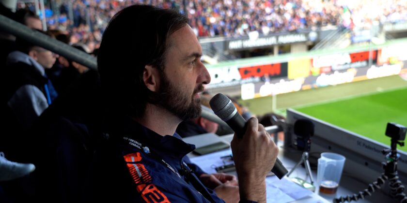 Ein Mann mit Bart und Kopfhörer hält ein Mikrofon und spricht in ein Stadion. Im Hintergrund sind Zuschauer und ein Fußballfeld zu sehen.