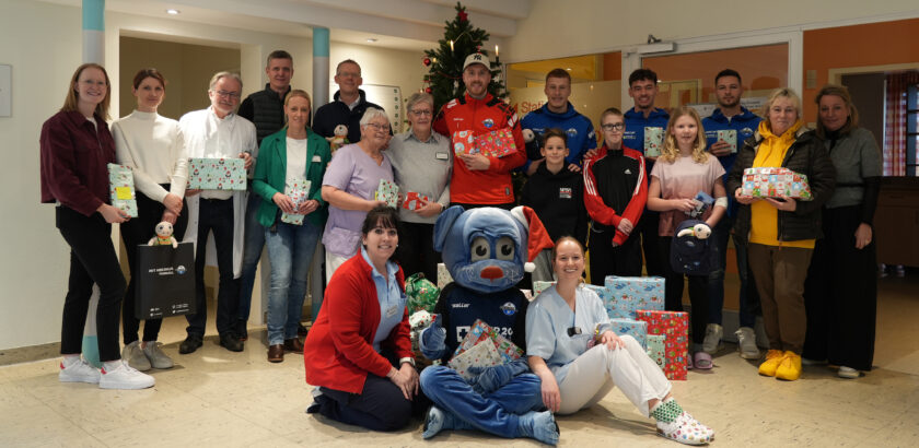 Eine Gruppe von Menschen, darunter medizinisches Personal und ein Maskottchen, hält eine Weihnachtsfeier in einem Krankenhaus ab. Sie halten Geschenke und Tüten in den Händen und stehen vor einem geschmückten Weihnachtsbaum. Die Atmosphäre ist festlich und freundlich.