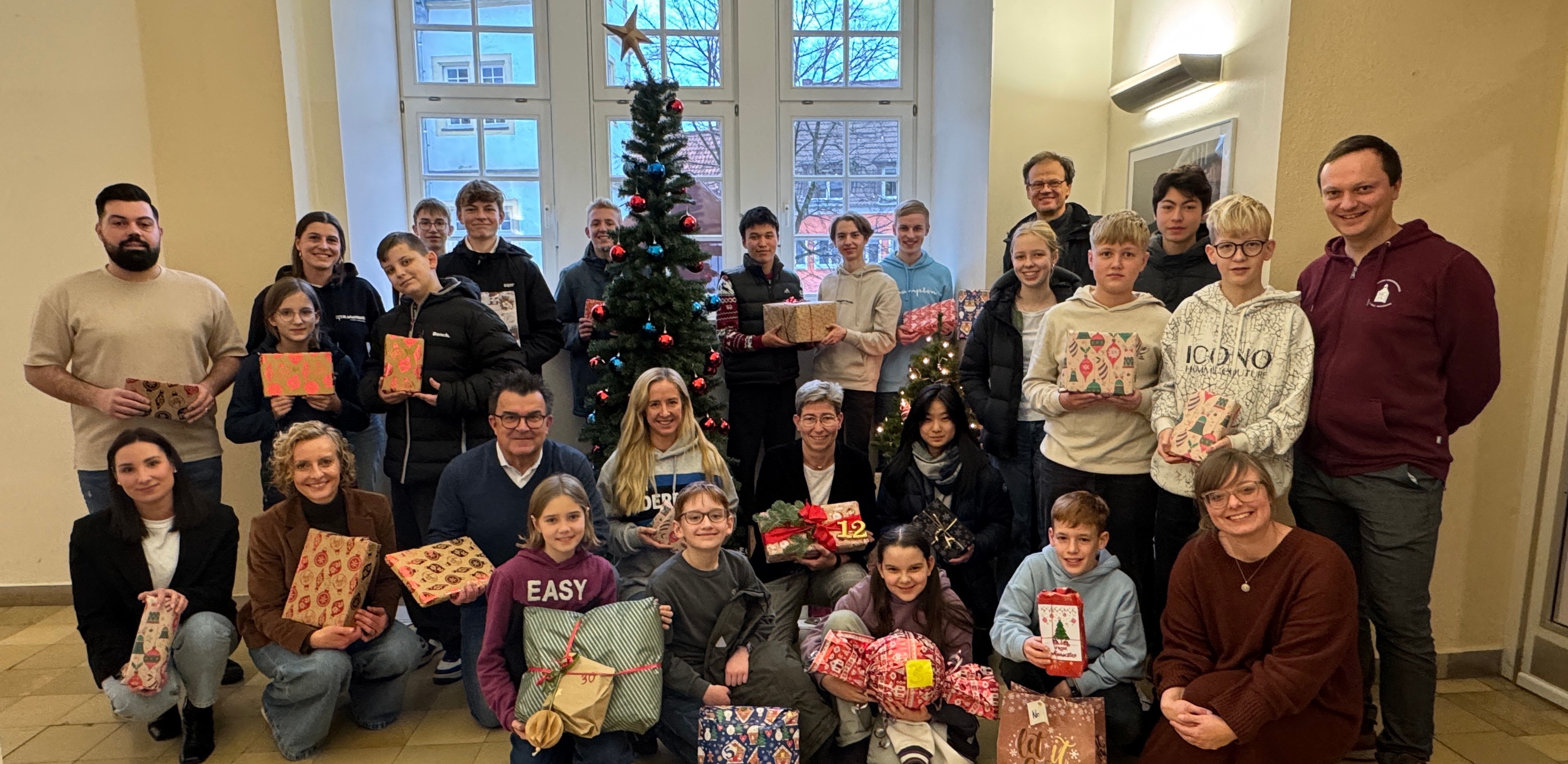 Eine Gruppe von Menschen, darunter Kinder und Erwachsene, steht in einem Raum mit großen Fenstern und einem geschmückten Weihnachtsbaum im Hintergrund. Alle halten bunt verpackte Geschenke in den Händen und lächeln in die Kamera. Die Atmosphäre wirkt festlich und fröhlich.