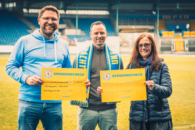 Drei Personen stehen auf einem Fußballfeld und halten jeweils einen Spendenscheck in den Händen. Sie lächeln in die Kamera. Die Person in der Mitte trägt einen Schal mit den Farben des Vereins, während die anderen beiden in Jacken gekleidet sind. Im Hintergrund sind die Sitzplätze des Stadions zu sehen.