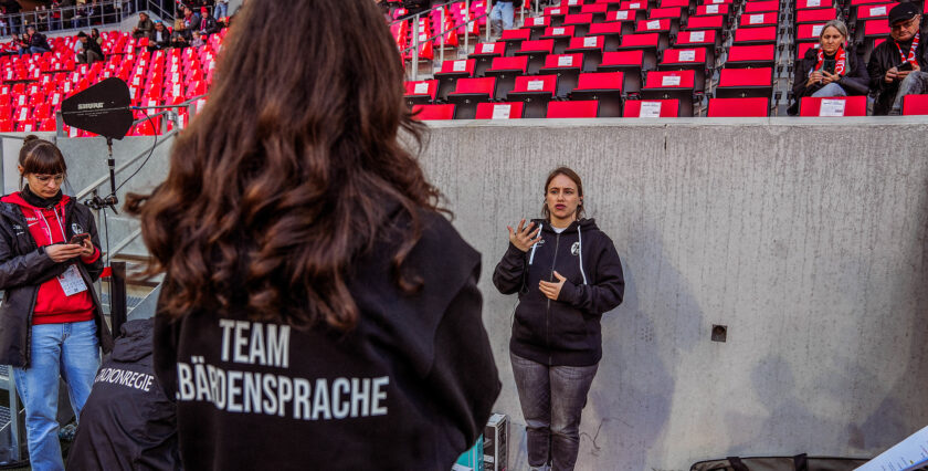 Eine Person mit einer Jacke, auf der "Team Gebärdensprache" steht, steht im Vordergrund und gebärdet. Im Hintergrund ist eine Person zu sehen, die gebärdet und sich in einem Stadion befindet. Die Sitzplätze im Stadion sind rot und teilweise besetzt. Eine weitere Person filmt die gebärdende Person mit einem Smartphone.
