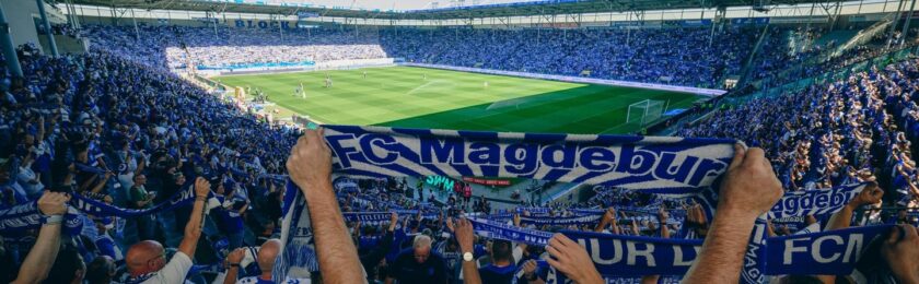 Ein Panoramabild eines vollen Fußballstadions mit jubelnden Fans. Die Fans halten große blaue und weiße Schals mit dem Aufdruck "FC Magdeburg" hoch. Das Spielfeld ist im Hintergrund zu sehen, und das Stadion ist mit vielen Zuschauern gefüllt, die die Atmosphäre eines spannenden Spiels widerspiegeln.