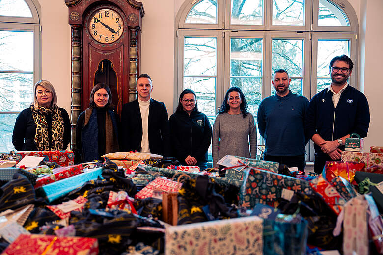 Eine Gruppe von sieben Personen steht lächelnd hinter einem Tisch, auf dem zahlreiche bunte Geschenktüten mit verschiedenen Mustern und Designs ausgebreitet sind. Im Hintergrund ist eine große Standuhr und ein Fenster mit Blick auf eine winterliche Landschaft zu sehen.