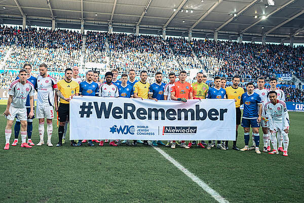 Eine Gruppe von Fußballspielern in verschiedenen Trikots steht auf dem Rasen eines Stadions und hält ein großes Banner mit der Aufschrift "#WeRemember". Im Hintergrund sind zahlreiche Zuschauer auf den Tribünen zu sehen. Das Bild erinnert an den Internationalen Tag des Gedenkens an die Opfer des Holocaust.