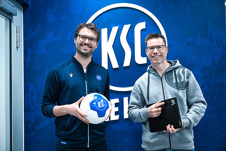 Zwei Männer stehen vor einem blauen Hintergrund mit dem Logo des KSC (Karlsruher SC). Der Mann links hält einen blauen und weißen Fußball in den Händen, während der Mann rechts ein schwarzes Tablet oder einen Ordner hält. Beide lächeln und tragen Kleidung mit dem KSC-Logo.