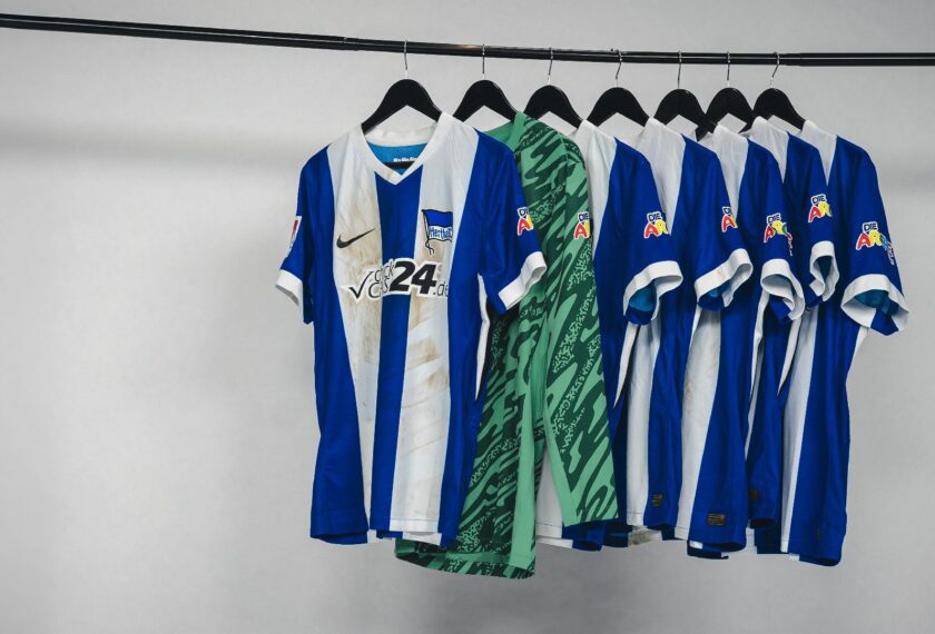 Mehrere Fußballtrikots des Bundesligisten Union Berlin hängen auf einer Stange. Die Trikots sind blau-weiß gestreift und tragen verschiedene Aufdrucke und Logos. Ein grünes Trikot mit Muster ist ebenfalls zu sehen. Die Trikots wurden offensichtlich getragen und sind Teil einer Versteigerungsaktion zugunsten der ARCHE.