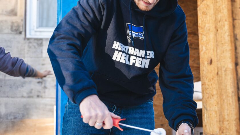 Ein Mensch in einem schwarzen Hoodie mit dem Logo des Hertha BSC und der Aufschrift "Herthaner Helfen" hält ein Werkzeug und beteiligt sich an einer Hilfsaktion.