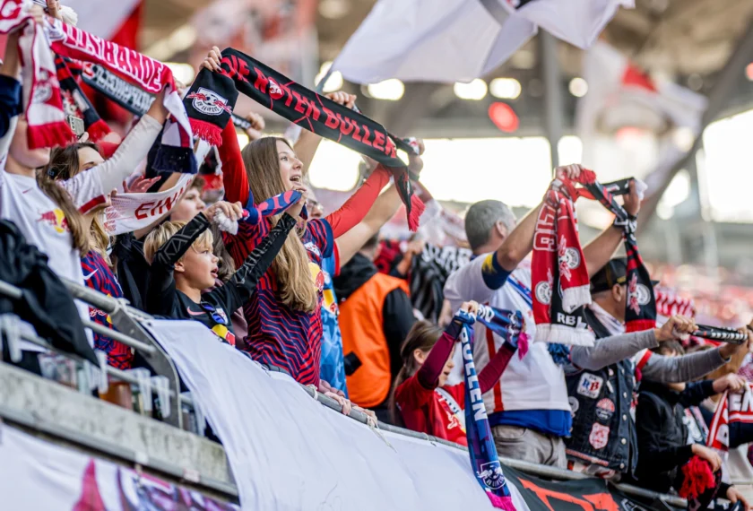 Fans im Stadion, die leidenschaftlich ihre Schals schwenken und die Mannschaft unterstützen. Die Fans tragen verschiedene Schals und Trikots in den Vereinsfarben und sind sichtbar begeistert.