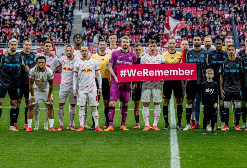 Zwei Fußballmannschaften stehen vor einem Spiel auf dem Rasen. Die Spieler der einen Mannschaft tragen weiße Trikots mit roten Streifen und die der anderen schwarzen Trikots mit blauem Logo. In der Mitte halten zwei Spieler ein Schild mit der Aufschrift "#WeRemember". Im Hintergrund sind zahlreiche Zuschauer auf den Tribünen zu sehen.