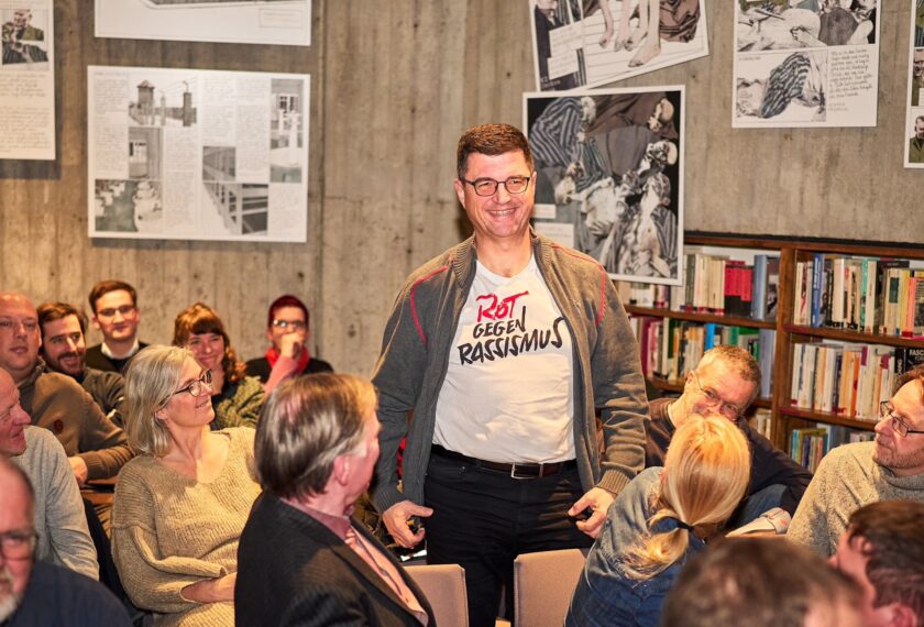 Ein Mann steht in einem Raum voller Menschen und trägt ein T-Shirt mit der Aufschrift "Rot gegen Rassismus". Er lächelt und scheint sich in einer Diskussion oder einem Workshop zu befinden. Im Hintergrund sind Comic-Bilder an der Wand und ein Bücherregal zu sehen.