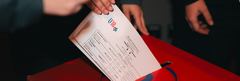 Eine Hand steckt einen ausgefüllten Wahlzettel in eine rote Wahlurne. Auf dem Wahlzettel ist das Logo der UEFA und der Schriftzug "U18-Wahl" zu sehen.