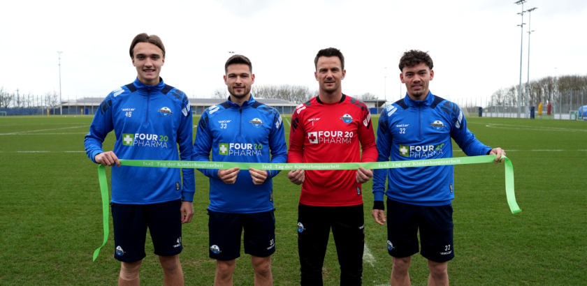 Vier Personen in Sportkleidung stehen auf einem Fußballfeld und halten gemeinsam ein grünes Band. Sie tragen T-Shirts mit dem Aufdruck "FOUR 20 PHARMA" und "Tag der Kinderhospizarbeit". Im Hintergrund sind Fußballtore und eine Tribüne zu sehen.