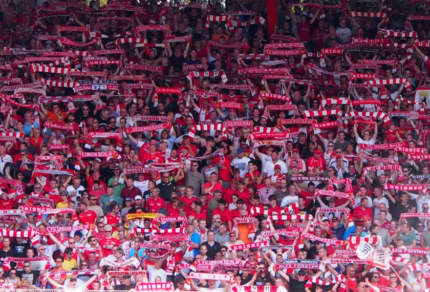 Eine große Menschenmenge in einem Stadion, die Fans des 1. FC Union Berlin sind. Die Fans tragen rote und weiße Schals und T-Shirts mit Aufschriften wie "Union Berlin", "FC Union Berlin" und "Eisern Union". Sie stehen dicht gedrängt und halten ihre Schals hoch, um ihre Unterstützung für das Team zu zeigen. Die Atmosphäre wirkt lebhaft und enthusiastisch.
