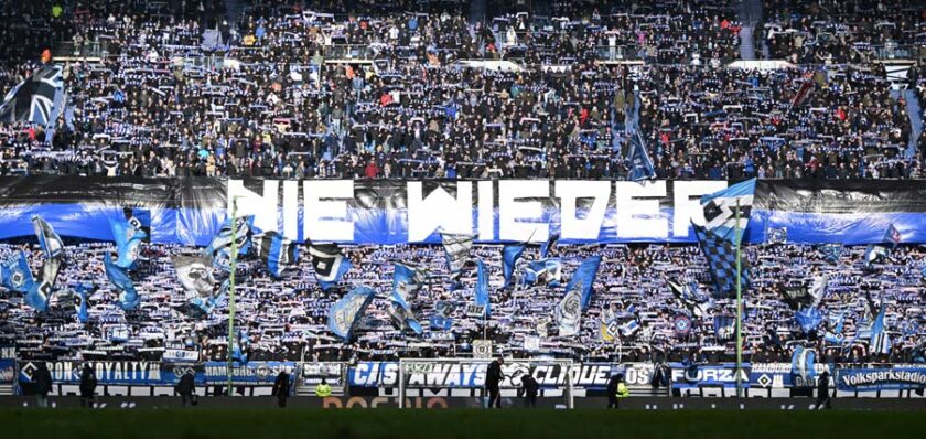 Eine große Menschenmenge in einem Fußballstadion, die ein großes Transparent mit der Aufschrift 'Nie Wieder' hält, umgeben von vielen blau-weißen Fahnen und Schals, die von den Fans geschwenkt werden.