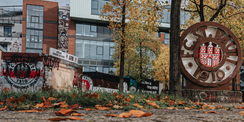 Ein Bild des Stadions des FC St. Pauli, das von Graffiti und Kunstwerken verziert ist. Im Vordergrund liegen herbstliche Blätter auf dem Boden, und im Hintergrund sind moderne Gebäude und Bäume mit herbstlichem Laub zu sehen.
