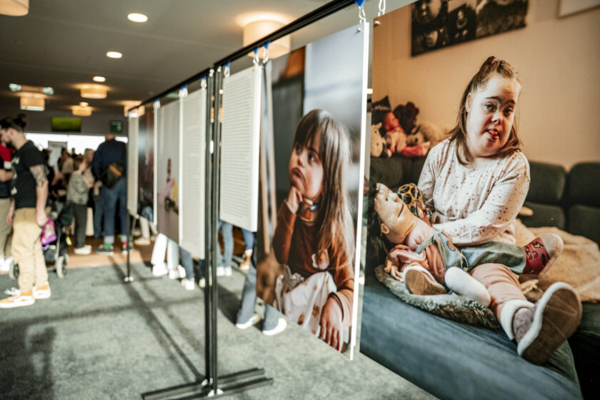 Ein großes Plakat zeigt ein Mädchen mit Down-Syndrom, das auf einem Sofa sitzt und ein Stofftier hält. Im Hintergrund sind Menschen in einem Raum mit Informationsständen zu sehen.