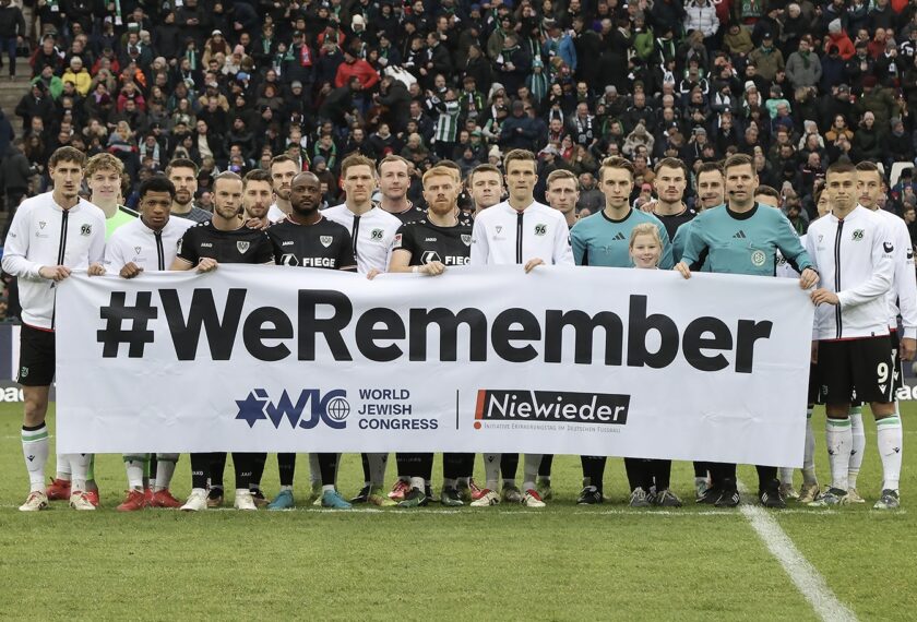 Ein Team von Fußballspielern und Schiedsrichtern steht auf dem Spielfeld und hält ein großes Banner mit der Aufschrift "#WeRemember". Im Hintergrund sind zahlreiche Zuschauer zu sehen. Das Banner zeigt auch die Logos des World Jewish Congress und der Initiative "Niewieder".