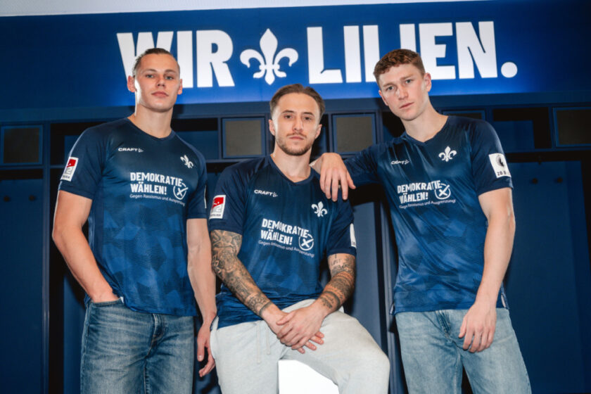 Drei Fußballspieler in blauen Trikots mit der Aufschrift "Demokratie wählen! Gegen Rassismus und Ausgrenzung" stehen vor einem blauen Hintergrund mit dem Schriftzug "Wir Lilien". Die Spieler posieren für ein Gruppenfoto, wobei einer von ihnen sitzt und die anderen beiden stehen.