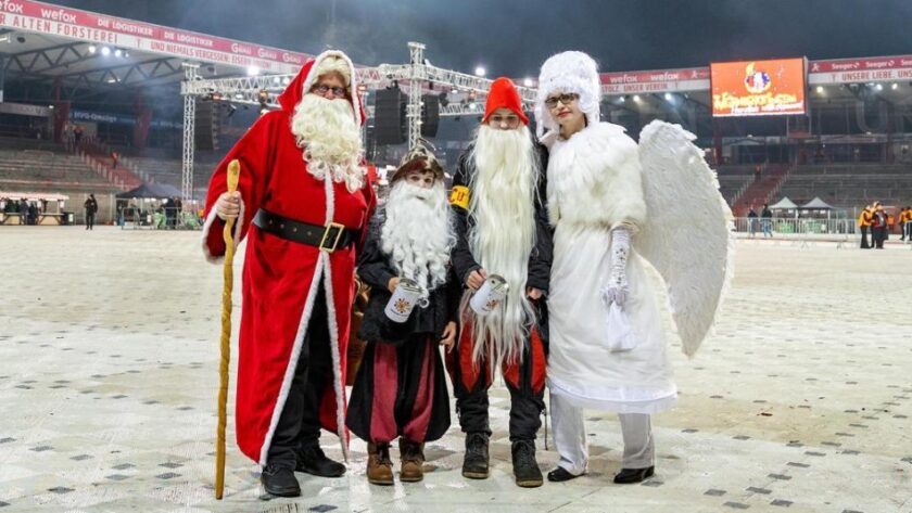 Vier Personen in Weihnachtskostümen stehen auf einem Fußballfeld. Von links nach rechts: Ein Mann als Weihnachtsmann mit rotem Mantel, weißem Bart und einem Stab, ein Kind als Weihnachtsmann mit rotem Mantel und weißem Bart, ein Mann als Weihnachtsmann mit rotem Mantel, weißem Bart und einem Becher in der Hand, und eine Frau als Engel mit weißem Gewand und Flügeln. Im Hintergrund sind Tribünen und eine Bühne zu sehen.