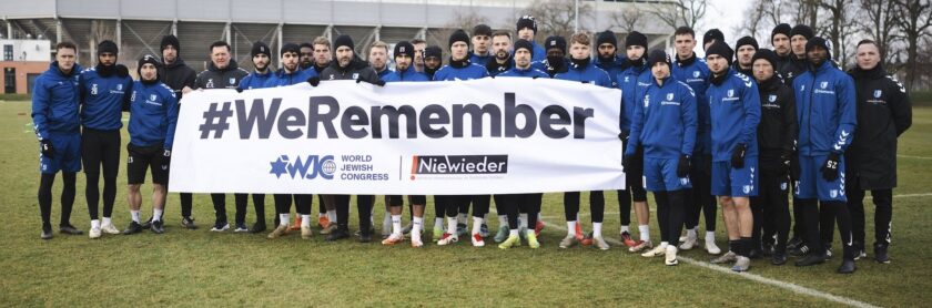 Eine Gruppe von Fußballspielern in blauen und schwarzen Trainingsanzügen steht auf einem Fußballfeld und hält ein großes Banner mit der Aufschrift "#WeRemember". Das Banner zeigt auch die Logos des World Jewish Congress und des Projekts "Niewieder". Die Spieler tragen Mützen und posieren für ein Gruppenfoto, um an historische Ereignisse zu erinnern.