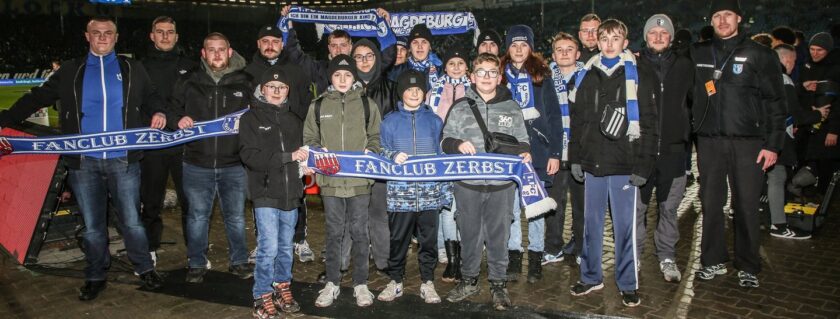 Eine Gruppe von Menschen, darunter Kinder und Erwachsene, steht vor einem Fußballstadion und hält ein blaues Banner mit der Aufschrift "Fanclub Zerbst". Einige der Anwesenden tragen Schals und Mützen in den Vereinsfarben. Die Gruppe scheint sich für ein Event oder eine Aktion im Stadion versammelt zu haben.