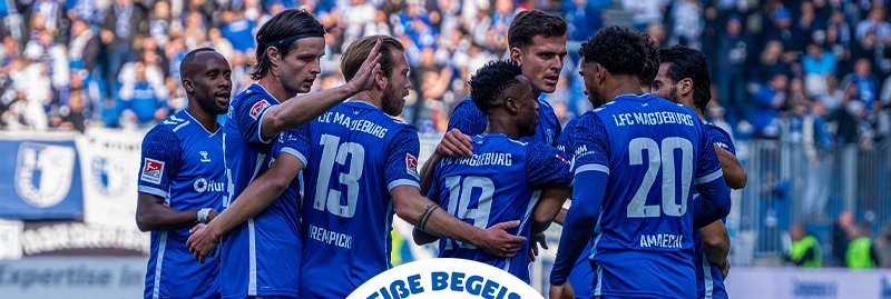 Eine Gruppe von Fußballspielern in blauen Trikots feiert gemeinsam auf dem Spielfeld. Die Spieler umarmen sich und zeigen ihre Freude über ein erfolgreiches Spiel. Im Hintergrund sind Zuschauer auf den Tribünen zu sehen.