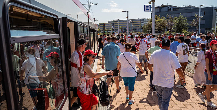 Eine Gruppe von Menschen steigt aus einem Zug aus und geht in Richtung eines Stadions. Die Menschen tragen verschiedene Kleidungsstücke, darunter T-Shirts, Shorts und Rucksäcke. Im Hintergrund sind moderne Gebäude und ein klarer blauer Himmel zu sehen.