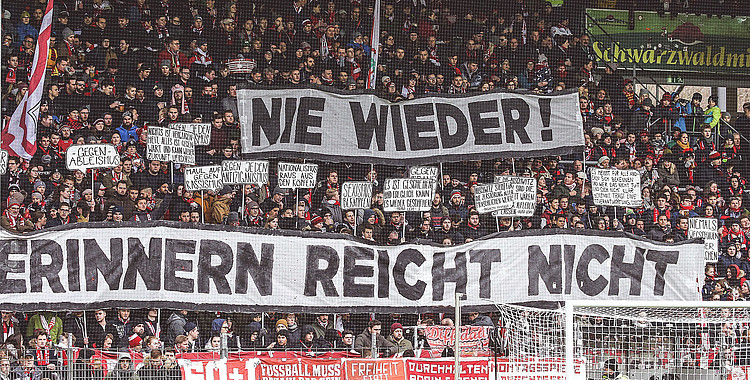 Eine große Gruppe von Fußballfans im Stadion, die ein Transparent mit der Aufschrift "NIE WIEDER! ERINNERN REICHT NICHT" hochhalten. Die Fans halten zusätzlich zahlreiche kleine Schilder mit verschiedenen Botschaften in den Händen. Im Hintergrund ist ein Teil des Spielfelds und der Stadiontribüne zu sehen.