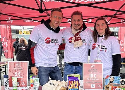 Drei Personen stehen lächelnd vor einem Stand mit verschiedenen Lebensmitteln. Sie tragen T-Shirts mit dem Aufdruck "Heimspiel gegen Armut" und "I ♥ Fußball". Der Stand ist mit einem roten Zelt bedeckt und scheint Teil einer Aktion zur Lebensmittelspende zu sein.