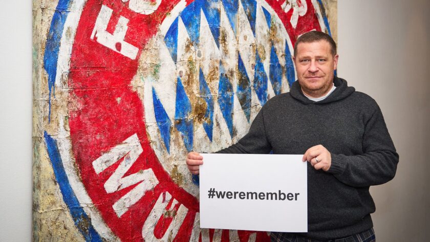 Ein Mann hält ein Schild mit der Aufschrift "#weremember" vor sich. Im Hintergrund ist ein Teil eines rot-blauen Schals mit dem Wort "Bayern" zu sehen.
