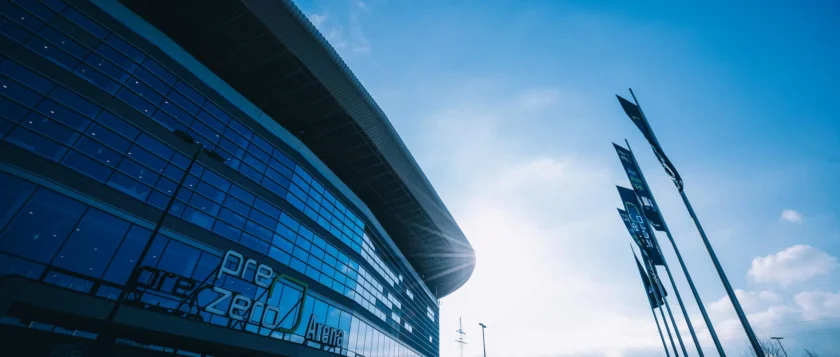 Moderne Arena mit der Aufschrift 'PreZero Arena' an der Fassade. Die Fassade besteht aus großen Glasfenstern und hat ein überhängendes Dach. Im Vordergrund sind mehrere Flaggenmasten mit Werbebannern zu sehen. Der Himmel ist blau mit einigen Wolken.