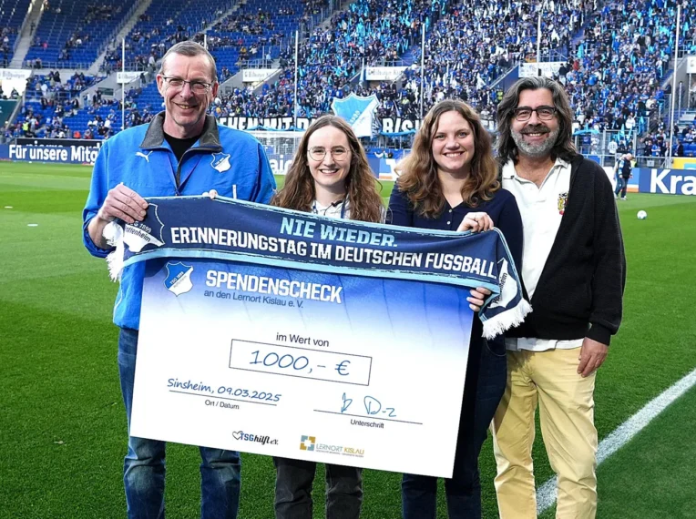 Vier Personen halten auf einem Fußballfeld ein großes blaues Tuch mit der Aufschrift "Nie wieder Erinnerungstag im Deutschen Fußball Spendenscheck an den Lemort Kislau e.V. im Wert von 1000,- €" und einem Logo. Im Hintergrund sind Zuschauer auf den Tribünen zu sehen.