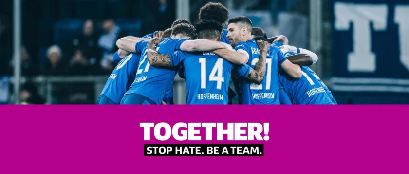 Eine Gruppe von Fußballspielern des TSG Hoffenheim feiert zusammen auf dem Spielfeld. Sie tragen blaue Trikots mit der Aufschrift "Hoffenheim" und umarmen sich. Im Vordergrund steht der Slogan "TOGETHER! STOP HATE. BE A TEAM." auf einem pinken Hintergrund.