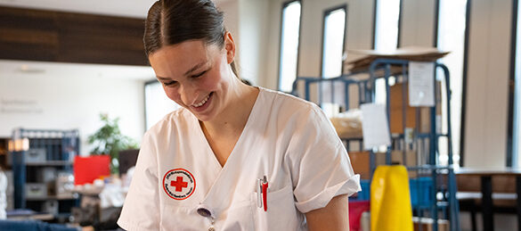 Eine lächelnde Frau in weißer Kleidung mit einem roten Kreuz-Emblem auf der Brust steht in einem Raum, der wie eine Blutspendeeinrichtung aussieht. Sie scheint sich auf eine Aufgabe zu konzentrieren, möglicherweise im Zusammenhang mit der Organisation oder Durchführung einer Blutspendeaktion.