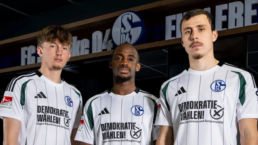 Drei Fußballspieler des FC Schalke 04 in weißen Trikots mit grünen Streifen und dem Aufdruck "Demokratie wählen! Gegen Rassismus und Ausgrenzung". Sie stehen vor einem Hintergrund mit dem Logo des Vereins und dem Schriftzug "VELTINS-ARENA".