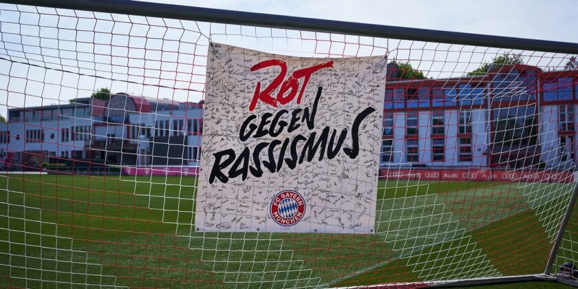 Ein Tor mit einem Banner, das die Aufschrift "Rot gegen Rassismus" trägt und mit vielen Unterschriften bedeckt ist. Im Hintergrund ist ein Fußballfeld mit einem Gebäude zu sehen.
