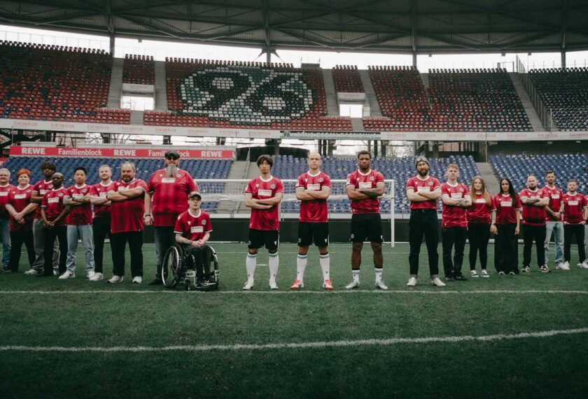 Gruppe von Menschen auf einem Fußballfeld im Stadion, darunter Spieler in roten Trikots und schwarzen Hosen sowie weitere Personen in Alltagskleidung. Im Hintergrund sind die Sitzplätze des Stadions mit der Aufschrift "REWE Familienblock" zu sehen. Eine Person im Rollstuhl ist Teil der Gruppe. Die Atmosphäre wirkt feierlich und organisiert, möglicherweise eine Ankündigung oder ein Gruppenfoto.