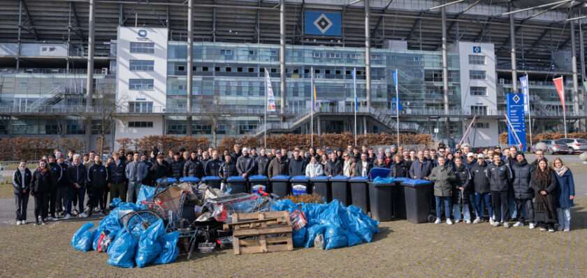 Eine große Gruppe von Menschen steht vor einem modernen Gebäude mit dem Logo des Hamburger SV. Im Vordergrund sind mehrere blaue Müllsäcke und andere Abfallmaterialien zu sehen, die auf einer Palette und dem Boden liegen. Die Menschen scheinen an einer Müllsammelaktion teilgenommen zu haben.