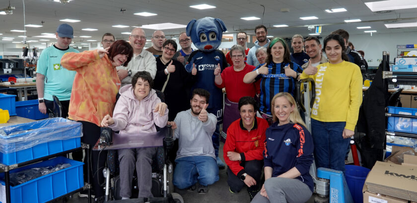 Eine Gruppe von Menschen in einem Lagerraum, darunter eine Person im Rollstuhl und eine Person mit einem Maskottchen-Kostüm. Alle lächeln und zeigen Daumen hoch, einige tragen T-Shirts mit Aufdrucken wie "saller" und "FOUR20 PHARMA". Im Hintergrund sind Regale und Kisten mit verschiedenen Gegenständen zu sehen.